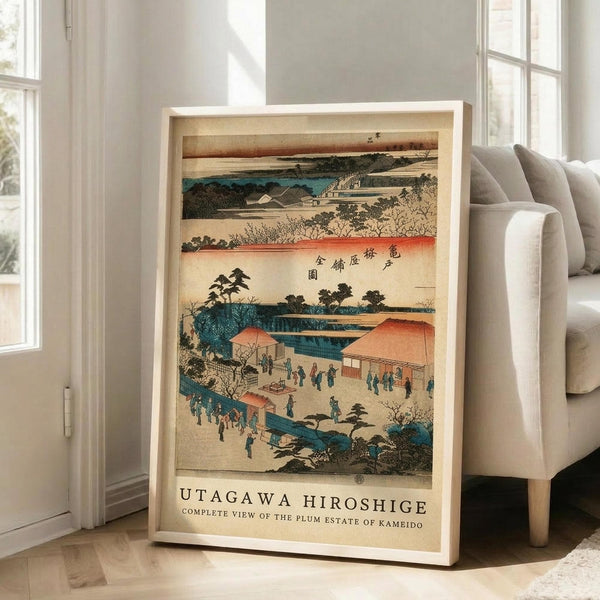 May include: Framed Japanese woodblock print showing a scenic landscape with buildings, bridge, and small figures in traditional dress. Print features muted tones of teal, coral, and cream with Japanese text at top. Light wood frame leans against white wall next to beige upholstered sofa in bright, modern living room with large windows and natural light casting shadows on hardwood floors.