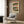 May include: A framed reproduction of a traditional Japanese woodblock print hanging on a wood-paneled wall above a beige upholstered armchair. The artwork features a landscape scene with blue water, orange buildings, small figures, and Japanese text, displayed in warm tones of blue, coral, and cream. A gold floor lamp and black side table with an open book complete the modern interior setting.
