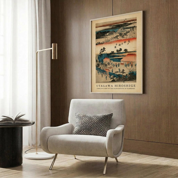 May include: A framed reproduction of a traditional Japanese woodblock print hanging on a wood-paneled wall above a beige upholstered armchair. The artwork features a landscape scene with blue water, orange buildings, small figures, and Japanese text, displayed in warm tones of blue, coral, and cream. A gold floor lamp and black side table with an open book complete the modern interior setting.