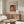 May include: A framed art print of Renoir's woman sewing displayed on a wooden sideboard in a minimalist living room. The artwork shows a woman in white clothing against warm orange tones. Modern furniture includes a cream curved chair, glass coffee table, and brass floor lamp. Neutral walls and natural lighting create a serene atmosphere.