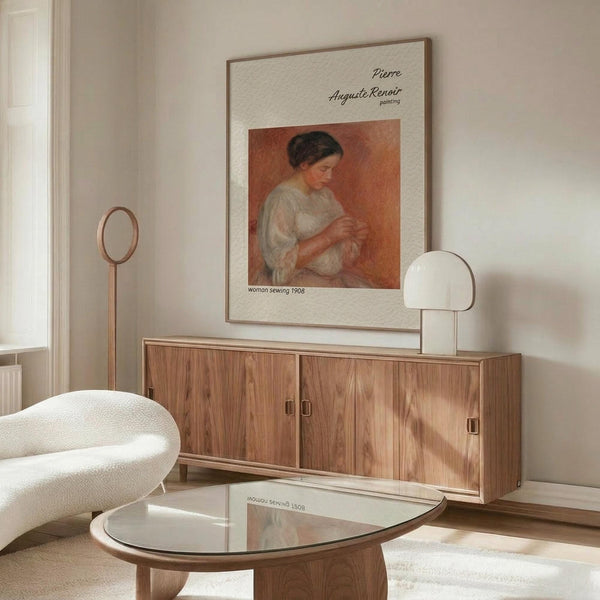 May include: A framed art print of Renoir's woman sewing displayed on a wooden sideboard in a minimalist living room. The artwork shows a woman in white clothing against warm orange tones. Modern furniture includes a cream curved chair, glass coffee table, and brass floor lamp. Neutral walls and natural lighting create a serene atmosphere.