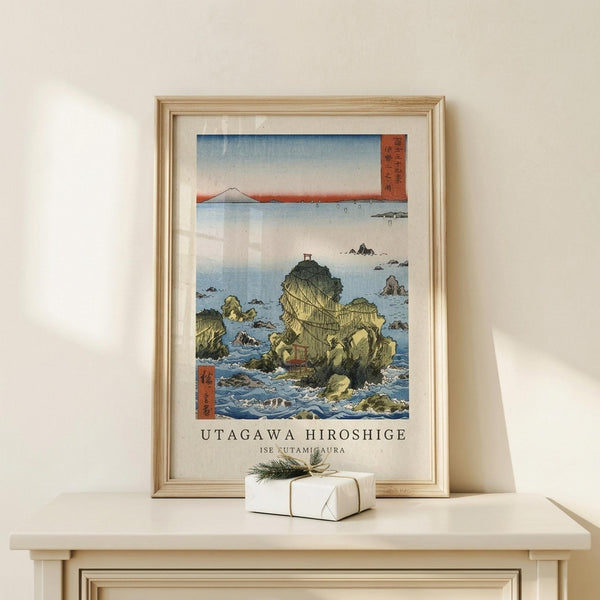 May include: A framed Japanese woodblock print displayed on a light wood console table against a cream wall. The artwork depicts rocky coastal islands in blue-green waters with Mount Fuji visible in the distance under a pale sky. A small white gift box with twine and evergreen sprig sits beside the natural wood frame. Soft natural lighting creates gentle shadows on the minimalist setting.