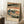 May include: A framed vintage Japanese woodblock print featuring a scenic landscape with traditional buildings, a winding blue river, small figures in robes, trees, and mountains under a peachy sky with Japanese characters, leaning against a white wall next to a wooden side table with coffee cup and saucer on a textured white rug.