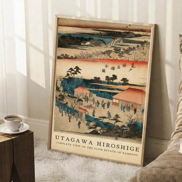 May include: A framed vintage Japanese woodblock print featuring a scenic landscape with traditional buildings, a winding blue river, small figures in robes, trees, and mountains under a peachy sky with Japanese characters, leaning against a white wall next to a wooden side table with coffee cup and saucer on a textured white rug.