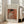 May include: Framed art print leaning against white wall next to beige upholstered chair in bright living room. Print features Pierre-Auguste Renoir painting from 1908 showing woman in white dress sewing against warm orange background. Light wood frame with white matting, soft natural lighting from large windows.