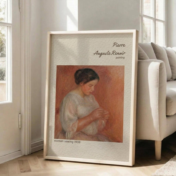 May include: Framed art print leaning against white wall next to beige upholstered chair in bright living room. Print features Pierre-Auguste Renoir painting from 1908 showing woman in white dress sewing against warm orange background. Light wood frame with white matting, soft natural lighting from large windows.
