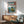 May include: Japanese woodblock print artwork displayed on white wall above wooden credenza, featuring rocky coastal landscape with small structures on cliffs, blue ocean waters, and Mount Fuji silhouette in distance under gradient sky. Print has traditional red Japanese text cartouches and warm wood frame. Scene includes mid-century modern sage green upholstered chair with wooden arms, white ceramic table lamp, and circular wood coffee table in sunlit room.