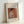May include: Framed art print showing Pierre Auguste Renoir's 1908 painting of a woman sewing, displayed in a light wooden frame against a white wall. The artwork features warm orange and peach tones with a woman in white clothing looking down at her needlework. Print has white matting with black text identifying the artist and artwork title.