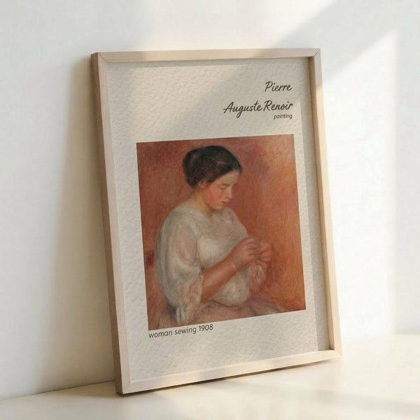 May include: Framed art print showing Pierre Auguste Renoir's 1908 painting of a woman sewing, displayed in a light wooden frame against a white wall. The artwork features warm orange and peach tones with a woman in white clothing looking down at her needlework. Print has white matting with black text identifying the artist and artwork title.