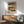 May include: Vintage Japanese woodblock print artwork displayed in wooden frame on white wall above light wood sideboard with white ceramic lamp. Features traditional landscape scene with buildings, trees, and figures in muted colors of blue, orange, and cream. Mid-century modern green upholstered armchair with wooden frame positioned nearby, creating serene living room interior with natural lighting and shadows.