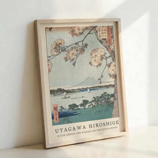 May include: Japanese woodblock print reproduction in light wooden frame leaning against white wall. Artwork shows cherry blossoms framing a scenic river view with Mount Fuji in distance, small boats on blue water, and green shoreline. Soft pastel colors with traditional ukiyo-e style. Text below reads "UTAGAWA HIROSHIGE" and "SUIJIN SHRINE AND MASSAKI ON THE SUMIDA RIVER" in black letters on white border.