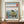 May include: Framed Japanese woodblock print reproduction displayed on wooden sideboard beside white ceramic vase and books. The artwork depicts rocky coastal formations surrounded by blue ocean waves, with Mount Fuji visible in the distant background under a gradient sky. Traditional red text cartouches appear in corners, and artist attribution text is printed below the image in a clean white mat.