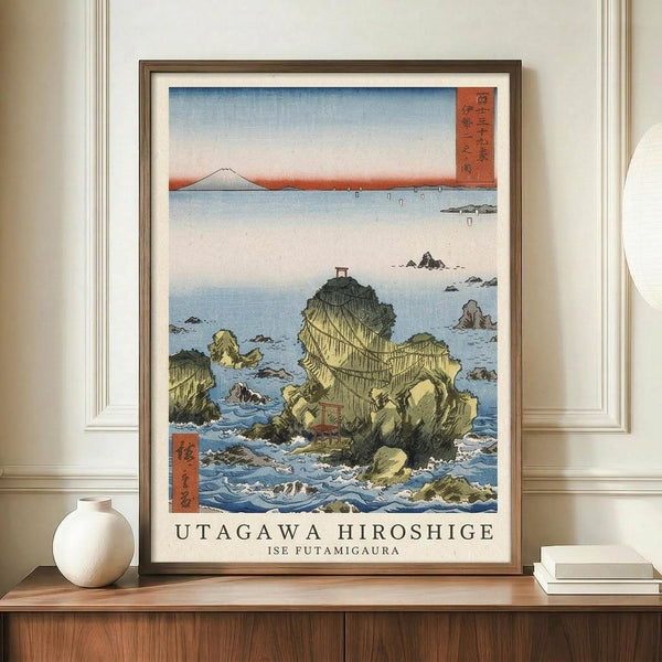May include: Framed Japanese woodblock print reproduction displayed on wooden sideboard beside white ceramic vase and books. The artwork depicts rocky coastal formations surrounded by blue ocean waves, with Mount Fuji visible in the distant background under a gradient sky. Traditional red text cartouches appear in corners, and artist attribution text is printed below the image in a clean white mat.