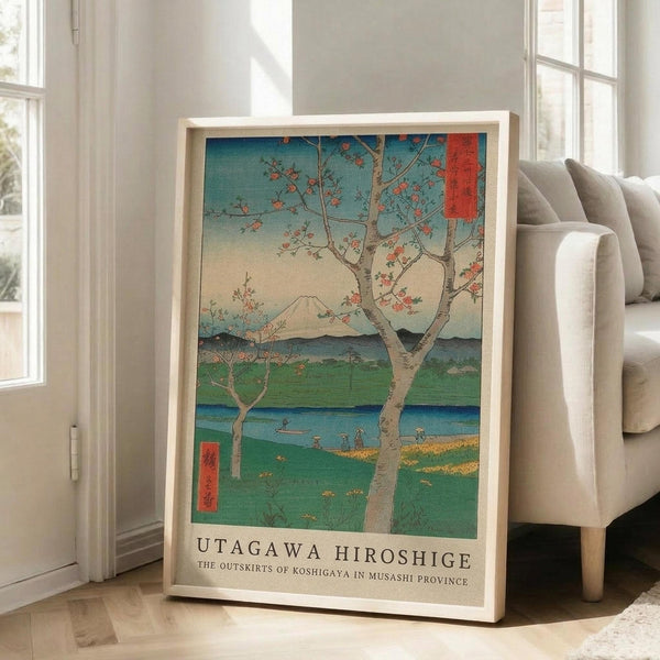 May include: Framed Japanese woodblock print reproduction leaning against white wall beside beige sofa. Artwork depicts cherry blossom tree in foreground with Mount Fuji in background, rendered in traditional ukiyo-e style with blue sky, green fields, and water. Print features red text cartouches and artist attribution at bottom in light wood frame.