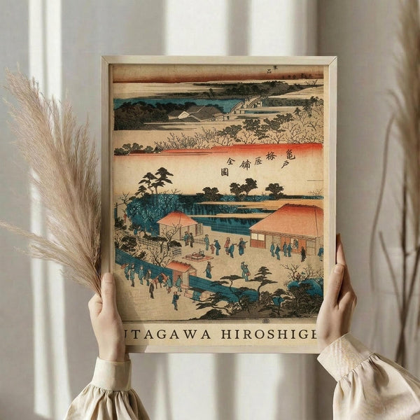 May include: Person holding framed Japanese woodblock print showing traditional landscape with buildings, people, and water in muted blues, oranges, and earth tones. Light wooden frame, displayed against white curtains with dried pampas grass visible on left side.
