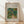 May include: Framed poster print of Pierre Auguste Renoir's impressionist painting showing two small figures walking along a winding path through lush green foliage and trees. The artwork features soft brushstrokes in greens, blues, yellows and coral tones. The print has a beige mat border with black text identifying the artist and title. Natural wood frame rests against white wall on light wooden floor with gentle sunlight shadows.