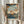 May include: Person in cream blouse holding framed Japanese woodblock print showing autumn maple branches with orange leaves overlooking a blue waterscape with mountains and traditional buildings. Light wood frame against white curtained background with dried pampas grass visible to the left.