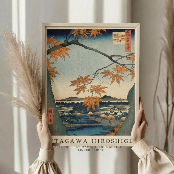 May include: Person in cream blouse holding framed Japanese woodblock print showing autumn maple branches with orange leaves overlooking a blue waterscape with mountains and traditional buildings. Light wood frame against white curtained background with dried pampas grass visible to the left.