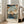 May include: Framed Japanese woodblock print featuring autumn maple branches with orange-red leaves overhanging a blue river landscape with mountains and traditional buildings. The artwork shows a serene water scene viewed through maple tree branches, displayed in a light wooden frame leaning against a white wall next to a beige upholstered chair in a bright, minimalist living room with large windows.