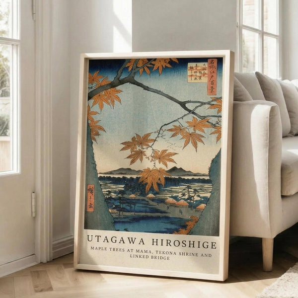 May include: Framed Japanese woodblock print featuring autumn maple branches with orange-red leaves overhanging a blue river landscape with mountains and traditional buildings. The artwork shows a serene water scene viewed through maple tree branches, displayed in a light wooden frame leaning against a white wall next to a beige upholstered chair in a bright, minimalist living room with large windows.