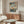 May include: Japanese woodblock print poster in light wood frame hanging above walnut credenza in modern living room. Artwork shows autumn maple branches with orange leaves overlooking blue water and mountains. White sculptural chair and glass-top coffee table visible. Warm natural lighting creates shadows on beige wall.