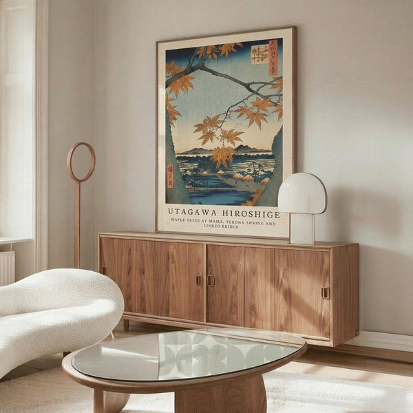 May include: Japanese woodblock print poster in light wood frame hanging above walnut credenza in modern living room. Artwork shows autumn maple branches with orange leaves overlooking blue water and mountains. White sculptural chair and glass-top coffee table visible. Warm natural lighting creates shadows on beige wall.