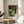 May include: Framed Japanese woodblock print featuring a dramatic waterfall cascading down white cliffs surrounded by red autumn maple trees and blue-green water. The vintage-style artwork displays traditional ukiyo-e aesthetics with Japanese text characters. The print sits on a light wood round side table next to a cream upholstered chair, with natural window light casting soft shadows and a potted plant visible in the corner of the neutral-toned room.