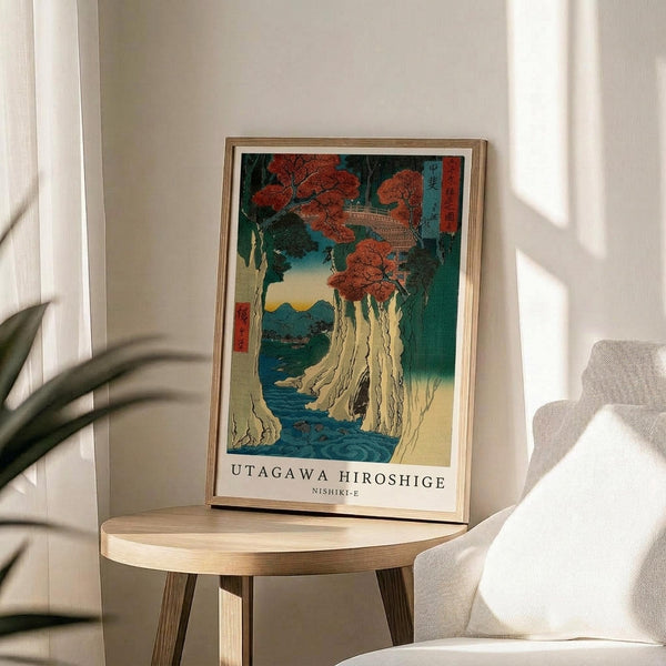 May include: Framed Japanese woodblock print featuring a dramatic waterfall cascading down white cliffs surrounded by red autumn maple trees and blue-green water. The vintage-style artwork displays traditional ukiyo-e aesthetics with Japanese text characters. The print sits on a light wood round side table next to a cream upholstered chair, with natural window light casting soft shadows and a potted plant visible in the corner of the neutral-toned room.