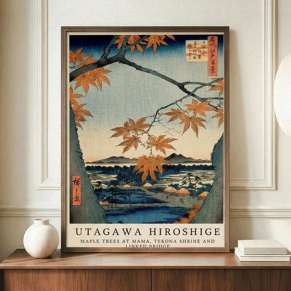May include: Framed Japanese woodblock print reproduction showing autumn maple branches with orange leaves framing a scenic view of mountains, water, and traditional buildings. The artwork features blue gradients in the sky, Japanese text cartouches, and earth tones throughout. Displayed on a wooden credenza beside a white ceramic vase and stacked books against cream-colored paneled walls.
