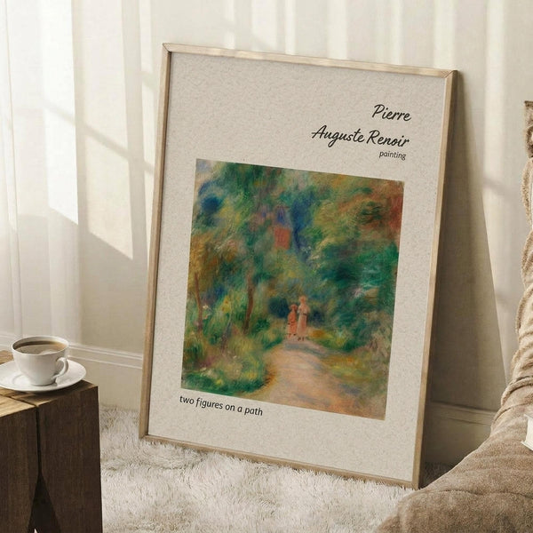 May include: Framed art print of Pierre-Auguste Renoir's impressionist painting showing two small figures walking on a winding path through lush green foliage with warm orange and blue accents. The print features a beige background with black text identifying the artist and artwork title. Light wood frame leans against white walls near a white coffee cup on wooden block and soft beige textiles on textured white rug.