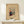 May include: Framed abstract art print featuring geometric shapes, lines, and vibrant colors in red, blue, yellow, and green on beige background. Wooden frame sits on light wood surface with small wrapped white gift box beside it. Museum exhibition poster style with artist name at bottom.