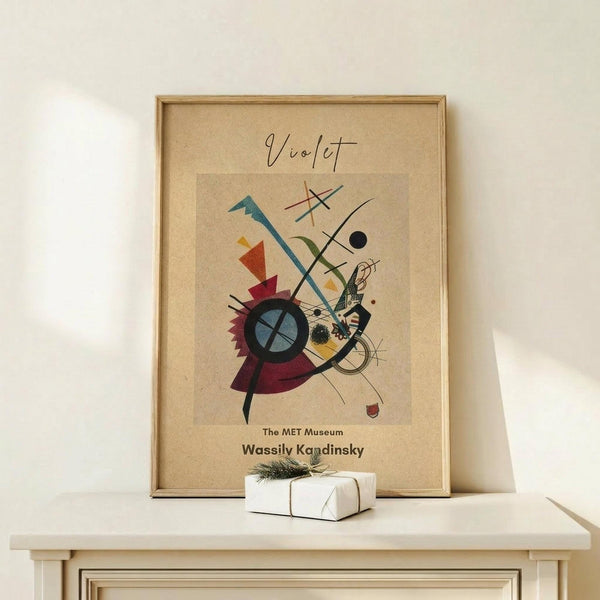 May include: Framed abstract art print featuring geometric shapes, lines, and vibrant colors in red, blue, yellow, and green on beige background. Wooden frame sits on light wood surface with small wrapped white gift box beside it. Museum exhibition poster style with artist name at bottom.