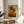 May include: Framed sunflower art print in warm golden and orange tones displayed on a round wooden side table next to a cream upholstered chair. The artwork shows three large sunflowers with detailed petals and leaves against a blue sky with sun, featuring Van Gogh's distinctive swirling brushstroke style. Natural lighting creates soft shadows across the neutral-toned room setting with a plant visible in the corner.