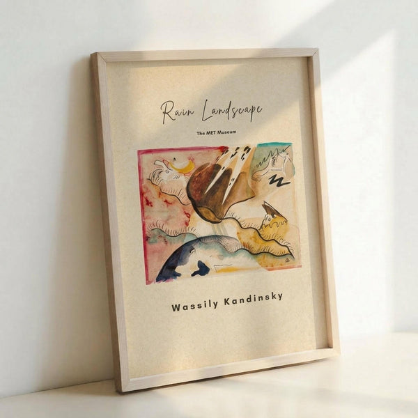 May include: Framed art print leaning against white wall showing abstract watercolor landscape with flowing organic shapes in browns, yellows, blues and reds. Cream-colored paper in light wood frame with "Rain Landscape," "The MET Museum," and "Wassily Kandinsky" text below the artwork. Soft natural lighting creates gentle shadows on the wall.