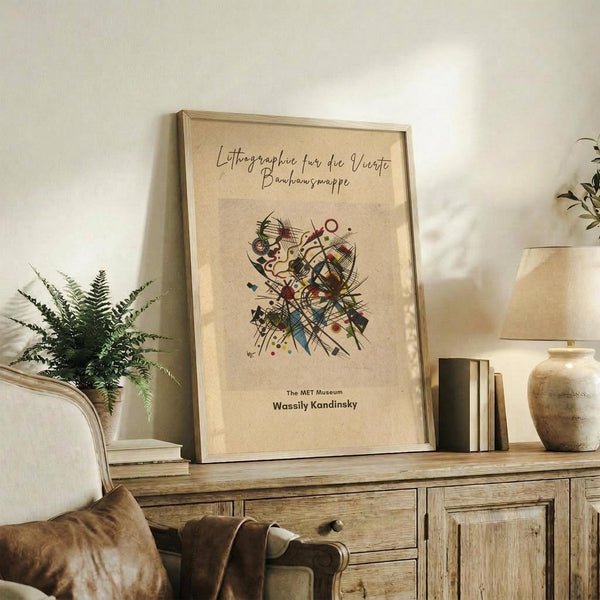 May include: Framed beige poster with abstract colorful design featuring geometric shapes, lines and circles in Kandinsky's style, displayed on rustic wooden sideboard next to fern plant, ceramic vase, and stacked books in neutral-toned living room setting with cream upholstered chair visible.