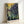 May include: Framed poster print of Van Gogh's Irises painting leaning against white wall. Artwork shows purple and blue iris flowers with flowing green stems and leaves against golden yellow background. Print displayed in light wood frame with white mat border. Van Gogh's signature visible at bottom. Characteristic post-impressionist brushwork and swirling patterns throughout composition.