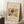 May include: A framed museum exhibition poster featuring Wassily Kandinsky's abstract watercolor artwork with flowing organic shapes in browns, blues, yellows, and reds. The cream-colored poster displays "Rain Landscape" and "The MET Museum" text above the artwork and "Wassily Kandinsky" below. The wooden-framed print leans against a white wall corner next to a beige blanket, with a coffee cup on a wooden block nearby on a textured cream rug.