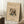 May include: Framed abstract art print leaning against white wall corner on textured white rug. Features colorful geometric composition with intersecting lines, circles, and angular shapes in blues, reds, yellows, and earth tones on cream background. German text at top, museum attribution at bottom. Light wood frame, positioned next to small wooden side table with white coffee cup and saucer. Soft natural lighting creates gentle shadows on wall.