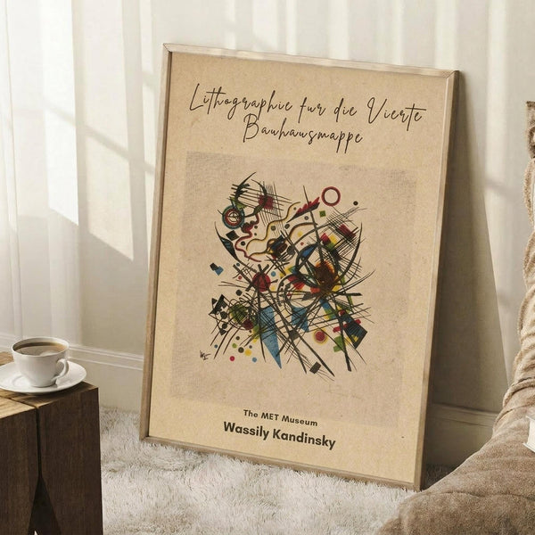 May include: Framed abstract art print leaning against white wall corner on textured white rug. Features colorful geometric composition with intersecting lines, circles, and angular shapes in blues, reds, yellows, and earth tones on cream background. German text at top, museum attribution at bottom. Light wood frame, positioned next to small wooden side table with white coffee cup and saucer. Soft natural lighting creates gentle shadows on wall.
