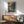 May include: Vincent van Gogh's Irises poster in wooden frame hanging on white wall above natural wood credenza with table lamp. Mid-century modern sage green armchair with wooden arms sits nearby on wooden floor with round coffee table. Sunlight creates dramatic shadows across the minimalist room setting.
