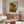 May include: Van Gogh sunflowers poster print in thin wooden frame hanging above walnut credenza in modern living room. Artwork shows three sunflowers with yellow petals and brown centers against blue sky and golden field. Room features cream curved chair, glass coffee table with wood base, brass floor lamp, and white table lamp on sideboard.