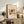May include: Framed art print featuring abstract watercolor artwork with flowing organic shapes in red, brown, blue, and yellow tones, displayed on rustic wooden sideboard alongside fern plant, books, and ceramic vase in neutral-toned living room setting with beige armchair visible