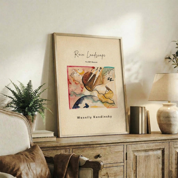 May include: Framed art print featuring abstract watercolor artwork with flowing organic shapes in red, brown, blue, and yellow tones, displayed on rustic wooden sideboard alongside fern plant, books, and ceramic vase in neutral-toned living room setting with beige armchair visible