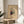 May include: Framed abstract art print featuring geometric shapes, lines, and vibrant colors including red, blue, yellow, and black on cream background. Wooden frame sits on light wood round side table next to beige upholstered chair. Soft natural lighting creates shadows across white walls. Green plant leaves visible in corner of minimalist, bright interior space.