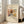 May include: Framed abstract art print leaning against white wall next to beige upholstered chair in bright living room. Artwork features swirling organic shapes in warm browns, yellows, blues and reds with flowing lines reminiscent of landscape forms. Light wooden frame with cream matting surrounds the colorful watercolor-style composition. Natural sunlight streams through tall windows creating soft shadows on white wooden floors.