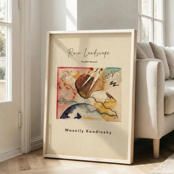 May include: Framed abstract art print leaning against white wall next to beige upholstered chair in bright living room. Artwork features swirling organic shapes in warm browns, yellows, blues and reds with flowing lines reminiscent of landscape forms. Light wooden frame with cream matting surrounds the colorful watercolor-style composition. Natural sunlight streams through tall windows creating soft shadows on white wooden floors.