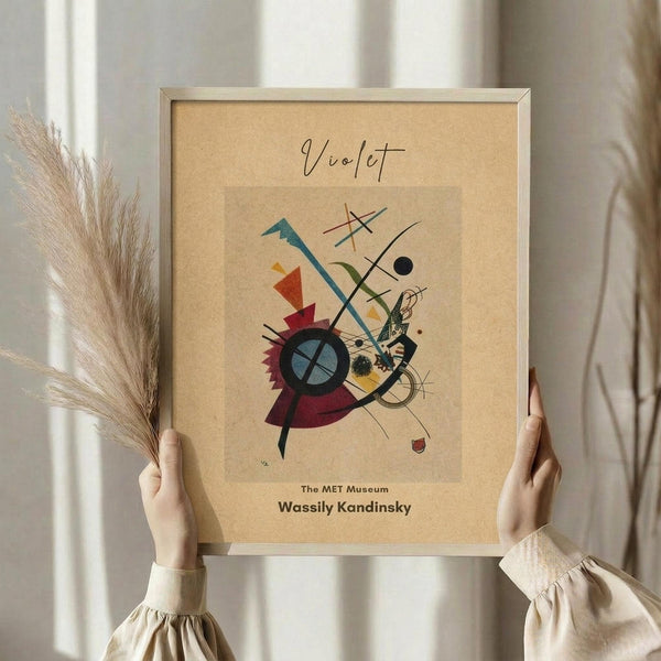May include: Hands holding framed abstract art print with geometric shapes, lines, and circles in red, blue, green, and yellow on beige background. Light wood frame, neutral bedroom setting with pampas grass and white curtains visible.