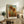 May include: Framed sunflower artwork in warm yellows and browns displayed on rustic wooden sideboard alongside fern plant, ceramic vase, and stacked books. The print shows three large sunflowers with detailed petals and leaves against blue sky and golden field background. Natural lighting creates soft shadows on white wall behind furniture.