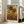 May include: Framed sunflower artwork in light wooden frame leaning against white wall next to beige upholstered chair. Print shows two large golden sunflowers with detailed petals and dark centers, green stems and leaves, set against blue sky with sun and golden wheat field below. Vintage-style poster with artist name at bottom.