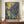 May include: Framed Van Gogh-inspired iris painting displayed on wooden sideboard against cream paneled wall. Artwork features purple and blue irises with flowing green stems against yellow-gold sky background in post-impressionist style. Brown frame, accompanied by white ceramic vase and stack of books on wood surface.
